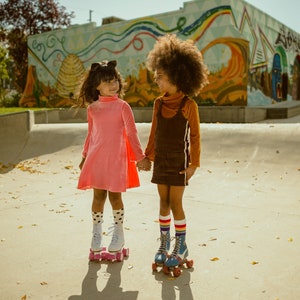 Op de afbeelding: Twee jonge meisjes in kleurrijke kleding en rollerskates houden elkaars hand vast en lachen naar elkaar voor een kleurrijk muurschildering.