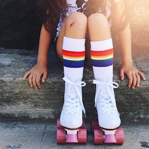 Op de afbeelding: Een persoon draagt witte rollerskates met roze wielen en regenboogkleurige gestreepte kniekousen. De persoon zit op een betonnen trede.