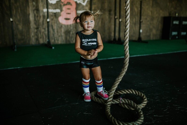 Op de afbeelding: Een jong meisje draagt een zwart tanktop met de tekst "Regulators" en regenboogkleurige gestreepte kniekousen en staat voor een dik touw.