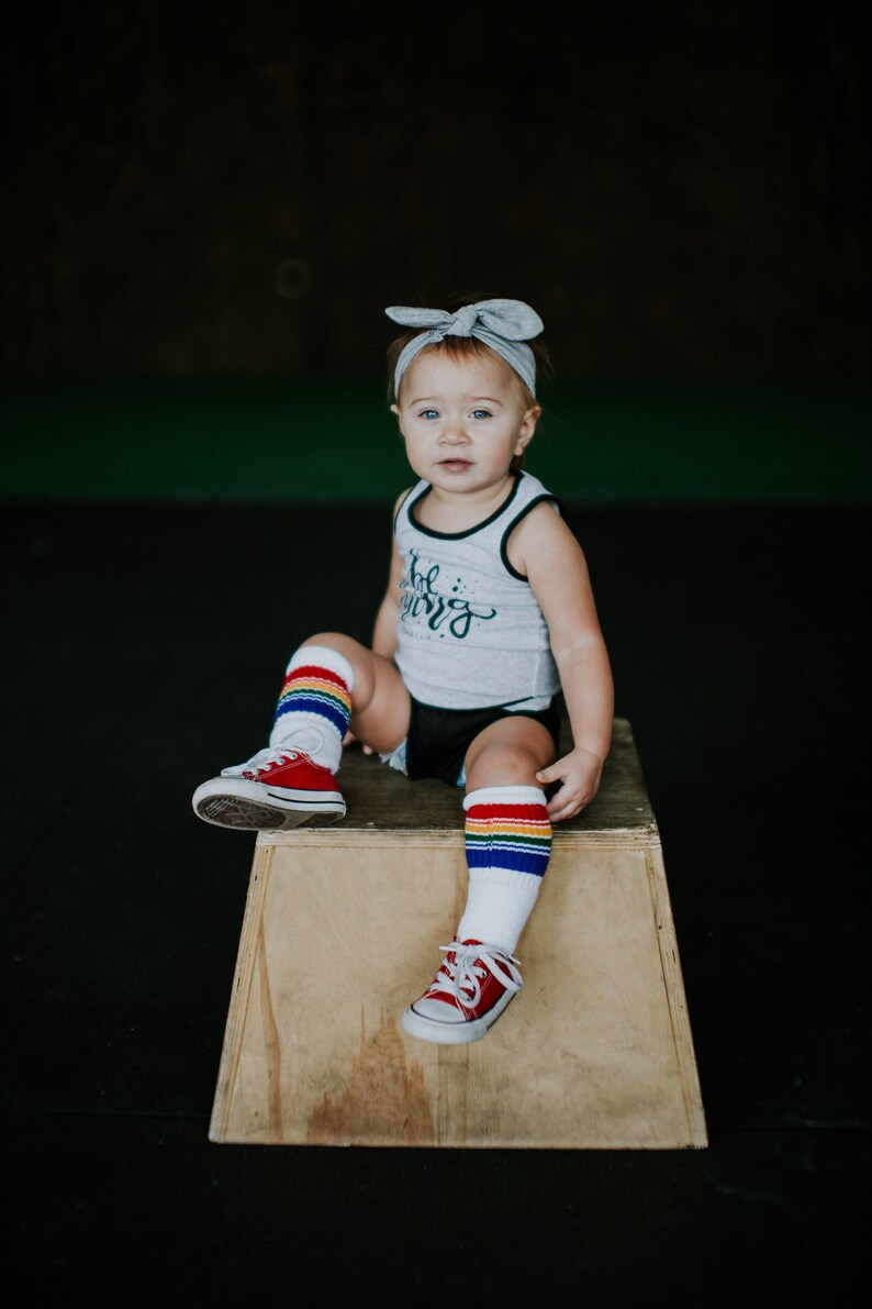 Op de afbeelding: Een jong kind draagt een grijs tanktop met de tekst "Little Warrior" erop gedrukt, zit op een houten kist. Het kind draagt regenboogkleurige, gestreepte kniekousen en rode sneakers.
