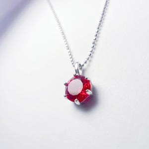 Op de afbeelding: Een zilveren ketting met een ronde, gefacetteerde, rode edelsteen hanger. De hanger is gezet in een zilveren bezel en hangt aan een fijne zilveren ketting. De edelsteen heeft een helder, reflecterend oppervlak.