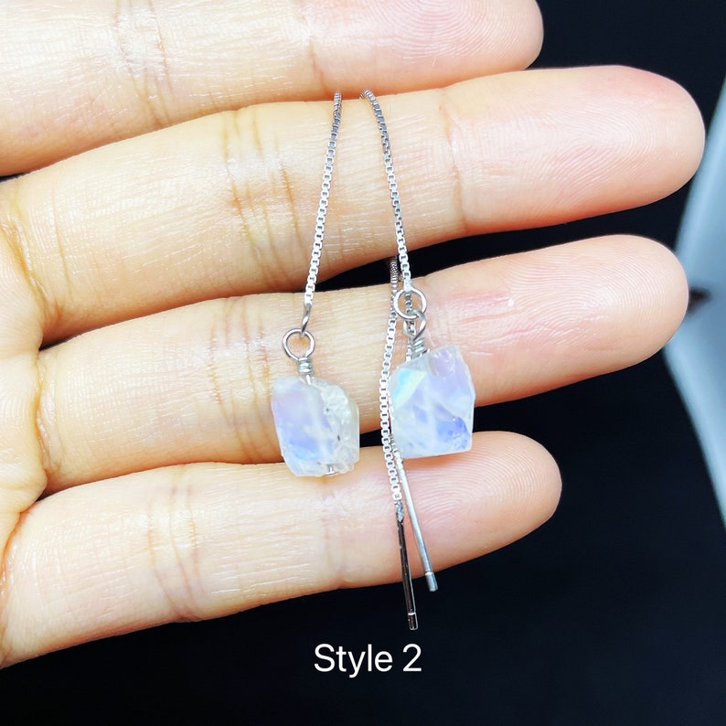 May include: A pair of silver dangle earrings with iridescent moonstone teardrop-shaped stones. The earrings are on a hand, and the text "Style 2" is visible below the earrings.