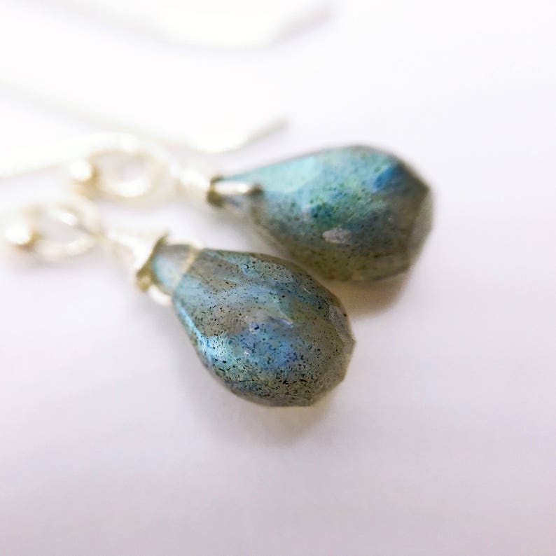 May include: A pair of silver dangle earrings with teardrop-shaped labradorite gemstones. The stones have a blue-green color with a shimmery, iridescent effect.