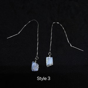 May include: A pair of silver dangle earrings with iridescent blue moonstone stones. The earrings are labeled "Style 3".