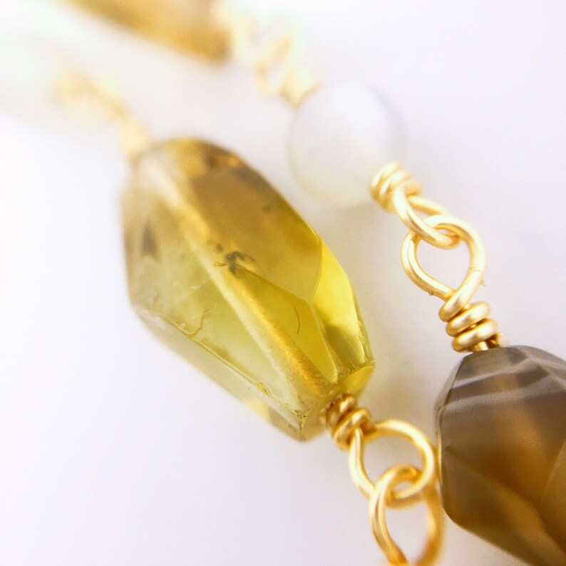 May include: A close-up of a gold chain with a yellow, faceted gemstone, a white gemstone, and a brown gemstone. The chain is made of wire and has a simple design.