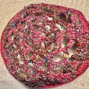 May include: A handmade, circular crochet item in shades of pink and silver, embellished with colorful sequins. The openwork design reveals a delicate pattern, creating a textured and decorative piece. The item appears to be a hair accessory or a decorative element.