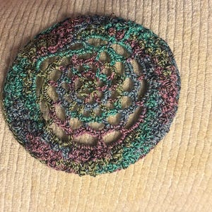 May include: A round, crocheted doily in shades of teal, purple, and silver. The openwork design features a central spiral pattern surrounded by a scalloped edge. The yarn has a subtle metallic sheen, adding a touch of sparkle.