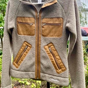 May include: A beige fleece jacket featuring a high collar, a full-length front zipper, and contrasting brown fabric panels on the chest and waist. The jacket includes two horizontal zippered chest pockets and two slanted zippered waist pockets.