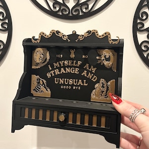 Puede incluir: Estante de madera negra con un diseño de tablero Ouija de color dorado. El tablero dice "I Myself Am Strange And Unusual" con las palabras "Yes", "No" y "Good Bye" impresas en el tablero. El estante tiene un pequeño cajón con detalles dorados.