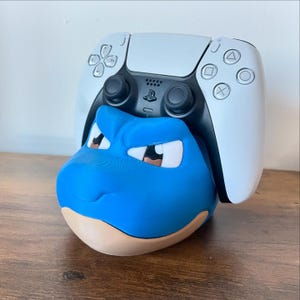 May include: A blue Blastoise-themed controller holder with a white PlayStation 5 controller resting on top. The holder is blue with a tan base and features the character's face. The controller is white and black.