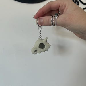 Puede incluir: Un llavero de cerámica blanca con forma de cráneo con una cuenca del ojo hueca. El llavero está unido a una cadena plateada.
