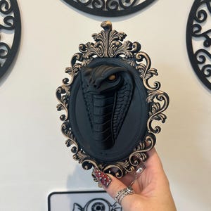 May include: A black cobra wall decoration in an ornate, oval frame with gold detailing. The cobra has detailed scales and an open hood, with an orange eye. The frame is held by a hand with rings and nail art. The sign below reads "DARK DIMENSIONS."