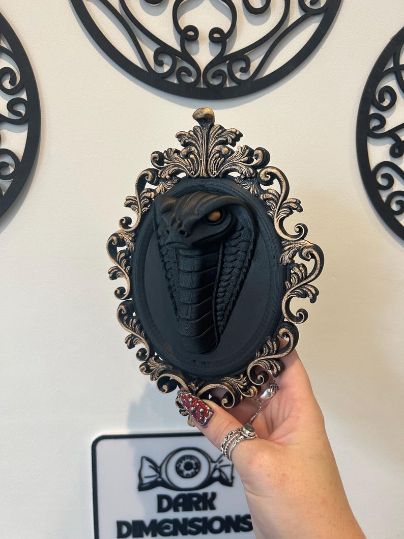 May include: A black cobra wall decoration in an ornate, oval frame with gold detailing. The cobra has detailed scales and an open hood, with an orange eye. The frame is held by a hand with rings and nail art. The sign below reads "DARK DIMENSIONS."