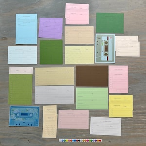 May include: A collection of colorful, blank index cards and two cassette tapes arranged on a wooden surface. The cards are in various pastel shades, with lines for writing. A strip of small, round, colorful stickers is at the bottom.