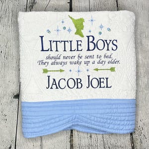 May include: White and blue baby quilt with a green Peter Pan silhouette and the text "LITTLE BOYS should never be sent to bed. They always wake up a day older. JACOB JOEL"