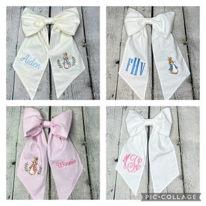 May include: Four white fabric hair bows with different embroidered designs. The top left bow has the name "Aiden" and a Peter Rabbit illustration. The top right bow has the monogram "FHV". The bottom left bow has the name "Winnie" and a Peter Rabbit illustration. The bottom right bow has the monogram "MJ".