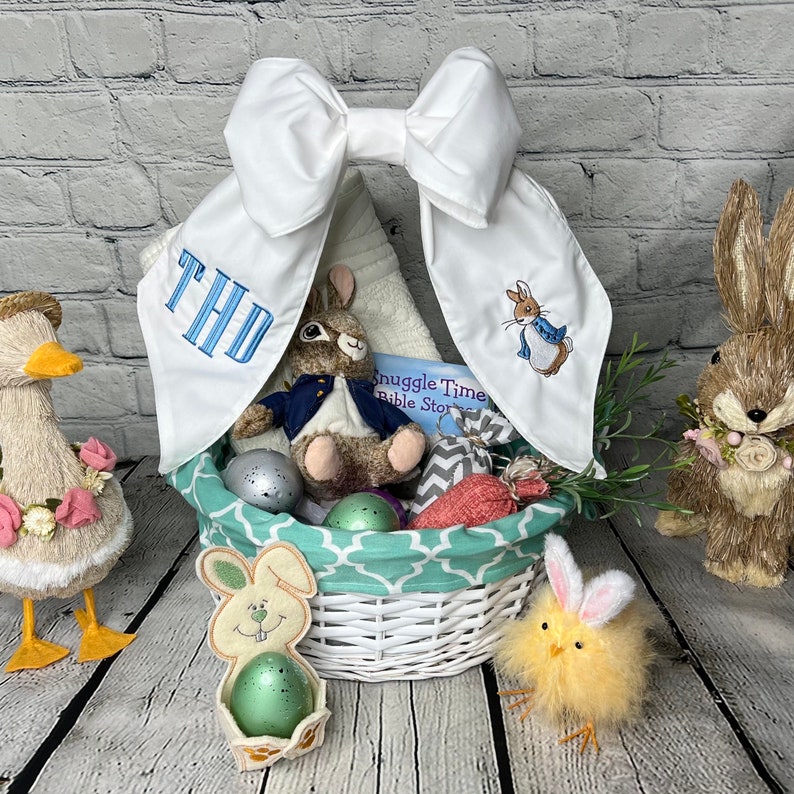 May include: A white wicker basket filled with Easter themed items, including a plush bunny, a book, and a stuffed duck. The basket is decorated with a white bow and a blue and green patterned liner. The basket is surrounded by Easter eggs and a small chick.