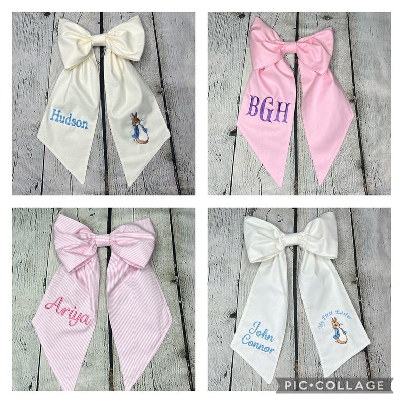 May include: Four white and pink fabric bows with different embroidered designs. The bows are tied in a large knot and have long tails. The designs include a bunny rabbit, the initials BGH, the name Ariya, and the name John Connor with the text "My First Easter".