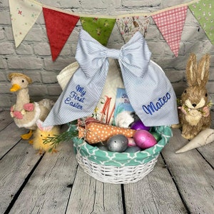 May include: A white wicker basket filled with Easter treats, including a plush carrot, plastic eggs, and a book. The basket is decorated with a blue and white striped bow with the text "My First Easter" and "Mateo" embroidered on it. A stuffed duck and a bunny are on either side of the basket.
