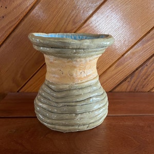 May include: A ceramic vase with a light brown and gray glaze. The vase has a wide mouth and a narrow neck, and it is decorated with a band of brown glaze around the middle. The vase is sitting on a wooden surface.