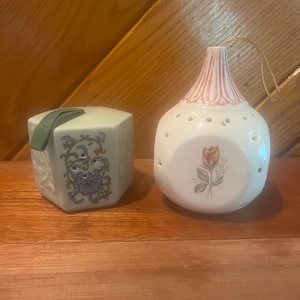 Puede incluir: Dos porta-potpourri de cerámica. Uno es de forma hexagonal con un diseño floral en verde, morado y rosa. El otro es redondo con una parte superior a rayas rosa y blanca y un diseño de una sola rosa en el centro.
