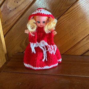 May include: A crocheted doll wearing a red dress with white trim and a white hooded cape. The doll has blonde hair and blue eyes.
