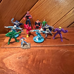 May include: A collection of 10 small plastic superhero figurines, including Superman, Batman, Spiderman, and The Flash. The figurines are in various poses and are displayed on a wooden surface.