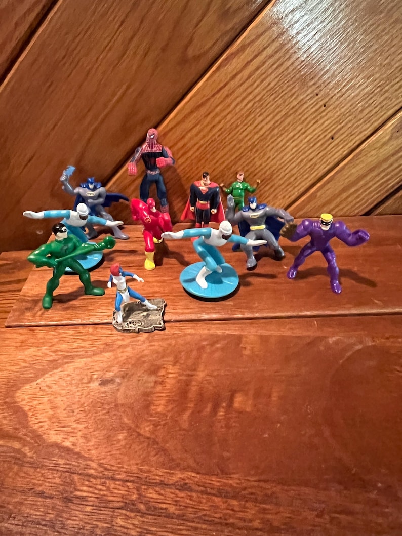 May include: A collection of 10 small plastic superhero figurines, including Superman, Batman, Spiderman, and The Flash. The figurines are in various poses and are displayed on a wooden surface.