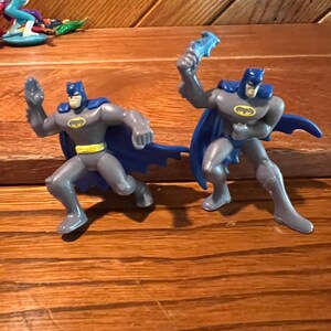 May include: Two gray plastic Batman action figures with blue capes and yellow belts. The figures are posed in a fighting stance.