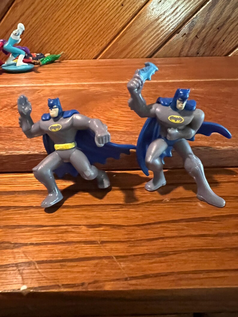 May include: Two gray plastic Batman action figures with blue capes and yellow belts. The figures are posed in a fighting stance.