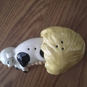May include: A ceramic salt and pepper shaker set featuring a black and white cow lying down with a yellow cabbage on top.