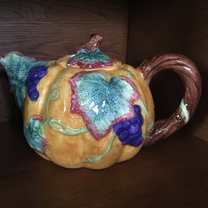 Op de afbeelding: Een keramische theepot in de vorm van een pompoen met een glanzende geel-oranje afwerking. De theepot is voorzien van verhoogde, handgeschilderde groene bladeren met roodachtige randen en trossen paarse druiven. Het heeft een bruin, getextureerd handvat.