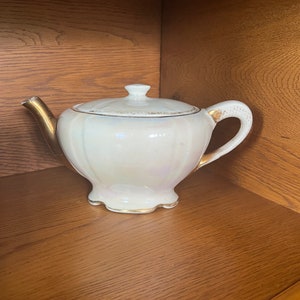 Puede incluir: Una tetera blanca con ribetes dorados y un acabado perlado. La tetera tiene una tapa y un asa.