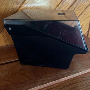 May include: Black plastic storage box with a clear plastic lid. The box has a beveled edge and a small rectangular label on the top.