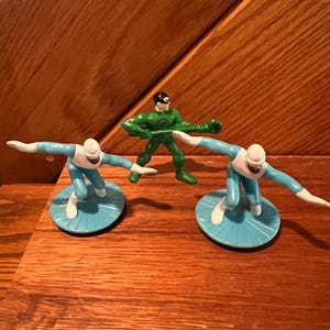 May include: Three plastic action figures, two in blue and white and one in green. The blue figures are on blue and white bases. The green figure is holding a green staff.