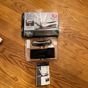 May include: A silver multi-tool with a black handle and a built-in LED flashlight. The tool is packaged in a clear plastic box with a black pouch and a printed instruction sheet. The packaging features the text "Pocket Tool" and "13 Functions Including Built-in LED Flashlight".