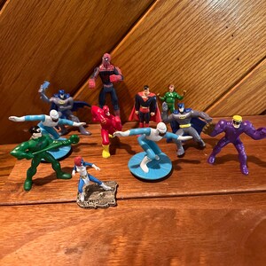 May include: A collection of superhero action figures, including Superman, Batman, Spiderman, The Flash, and others. The figures are made of plastic and are painted in various colors.