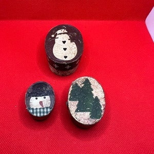 May include: Three small oval-shaped boxes with a Christmas theme. The boxes are decorated with a snowman, a Christmas tree, and a snowman's face.