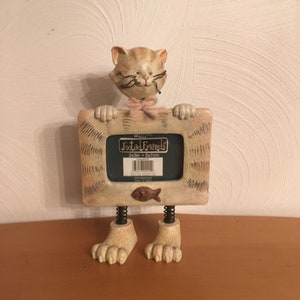 May include: A white cat figurine with black whiskers and glasses holding a picture frame. The frame is decorated with a striped pattern and has a brown label that reads "Footed Friends" and "2x3in - 5x7cm".