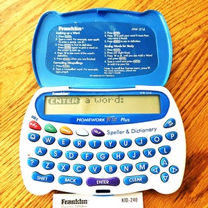 Peut inclure: Un dictionnaire et correcteur d'orthographe électronique Franklin Homework Wiz Plus blanc et bleu, modèle HW-216, avec un clavier et un couvercle ouvert affichant des instructions. Une petite carte blanche avec le logo Franklin est en dessous.