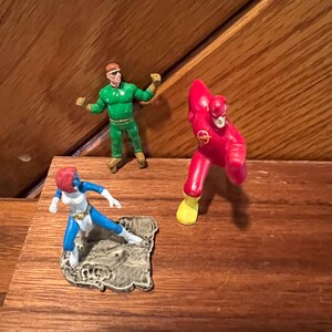 May include: Three small plastic action figures of superheroes. The figures are standing on a wooden surface. The figure on the left is blue and white, the figure in the center is green, and the figure on the right is red and yellow.