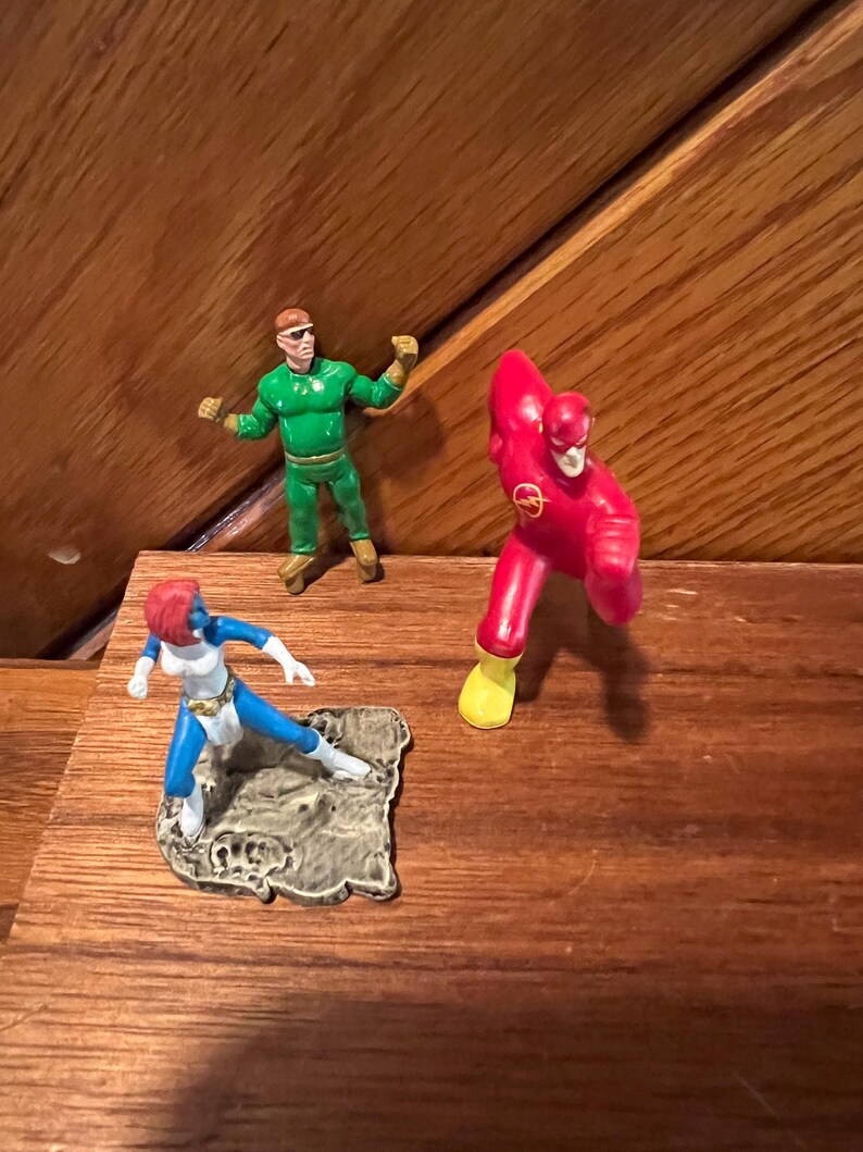 May include: Three small plastic action figures of superheroes. The figures are standing on a wooden surface. The figure on the left is blue and white, the figure in the center is green, and the figure on the right is red and yellow.