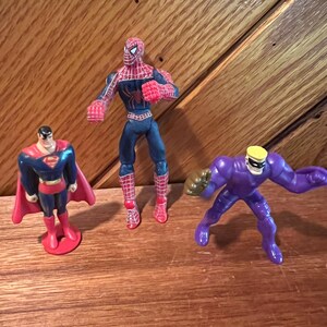 May include: Three superhero action figures: a red and blue Spiderman, a red and blue Superman, and a purple figure with a yellow helmet.