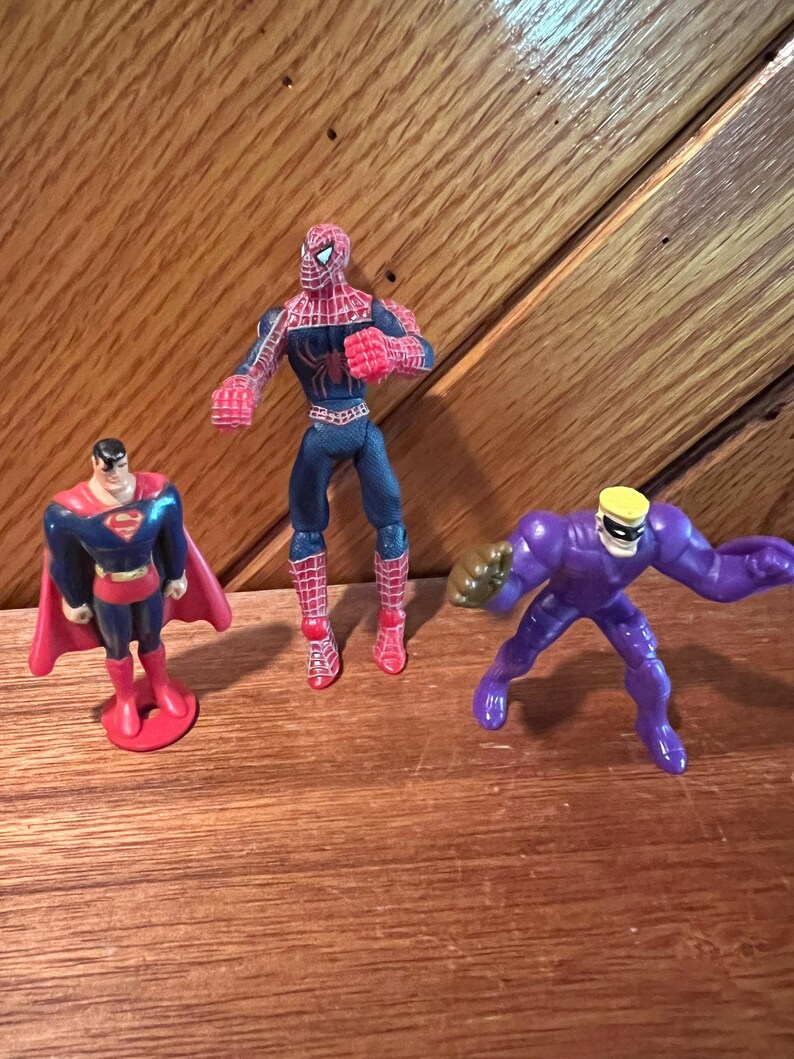 May include: Three superhero action figures: a red and blue Spiderman, a red and blue Superman, and a purple figure with a yellow helmet.