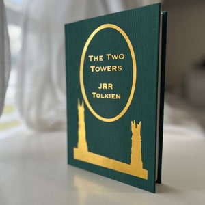 Może przedstawiać: Zielona książka w twardej oprawie z  złotym napisem "The Two Towers" autorstwa J.R.R. Tolkiena. Okładka przedstawia złotą sylwetkę dw&oacute;ch wież.
