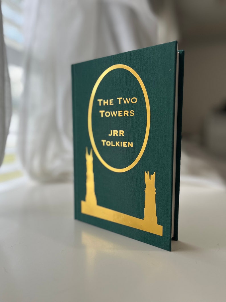 Może przedstawiać: Zielona książka w twardej oprawie z  złotym napisem "The Two Towers" autorstwa J.R.R. Tolkiena. Okładka przedstawia złotą sylwetkę dw&oacute;ch wież.