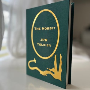 Może przedstawiać: Zielona książka w twardej oprawie ze złotym okręgiem wok&oacute;ł tytułu "Hobbit" autorstwa J.R.R. Tolkiena. Na okładce przedstawiono złotego smoka.
