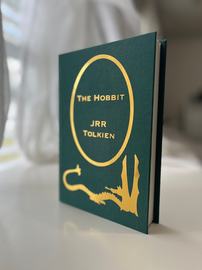 Może przedstawiać: Zielona książka w twardej oprawie ze złotym okręgiem wok&oacute;ł tytułu "Hobbit" autorstwa J.R.R. Tolkiena. Na okładce przedstawiono złotego smoka.