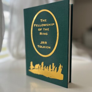 Może przedstawiać: Zielona książka w twardej oprawie z złotym okręgiem na okładce. Okrąg zawiera tekst "The Fellowship of the Ring" i "J.R.R. Tolkien". Na dole książki znajduje się złota sylwetka grupy ludzi idących.