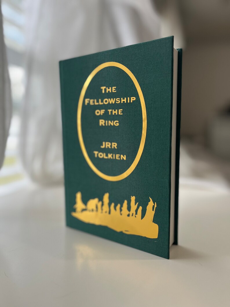 Może przedstawiać: Zielona książka w twardej oprawie z złotym okręgiem na okładce. Okrąg zawiera tekst "The Fellowship of the Ring" i "J.R.R. Tolkien". Na dole książki znajduje się złota sylwetka grupy ludzi idących.